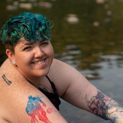 A non-binary person sits and relaxes in a shallow river. They are wearing blue jeans, a bikini top and sunglasses, and have bright blue hair.