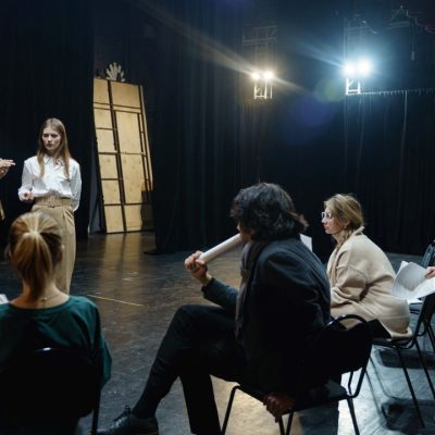 a group of people 3 sitting down and 3 standing up in a circle on a theater stage